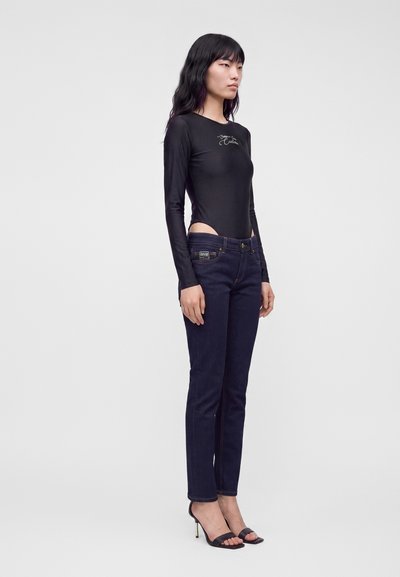 Femme debout portant un jean skinny taille haute foncé, un haut noir ajusté à manches longues avec des découpes aux hanches, et des sandales noires à talons hauts sur fond blanc.