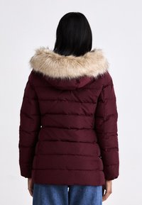 Burgundowa kurtka puchowa z kapturem z sztucznego futra, z pikowanym wzorem i dopasowanym krojem. Materiał wydaje się błyszczący i ocieplony.