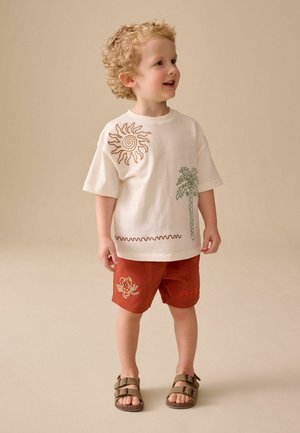 Lille barn med krøllet blondt hår iført en cremefarvet t-shirt med sol og palmetræ-design, rustfarvede shorts med krabbe-motiv og sandaler.