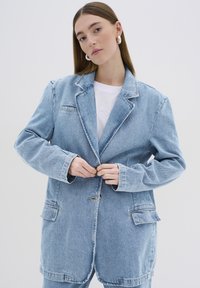 Svetlomodrý, oversize džínsový blejzer s jedným gombíkom, veľkými vreckami a uvoľneným strihom. Hladká textúra a klasický design klopy.