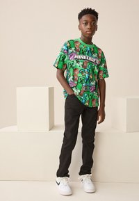 Groen T-shirt met korte mouwen met Minecraft-graphics, gecombineerd met een zwarte broek en witte sneakers. Het shirt heeft een opvallend grafisch logo.