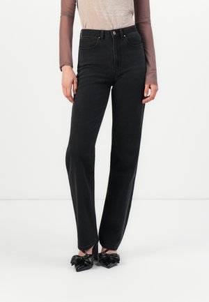 High-waisted black jeans in denim with a straight leg fit, paired with a long-sleeve sheer top and black bow-accented heels.