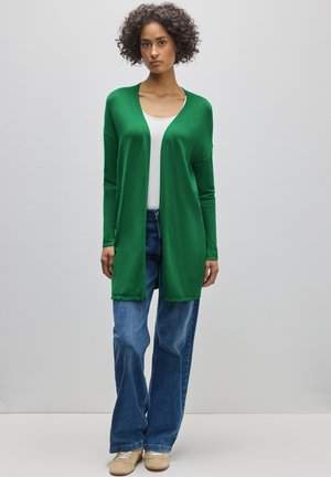 Nainen, jolla on kiharat hiukset ja päällään vihreä avoin neuletakki, valkoinen toppi, siniset leveälahkeiset farkut ja beiget tennarit seisomassa yksinkertaista seinää vasten.