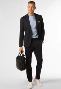 Dunkelblazer über einem hellblauen T-Shirt, kombiniert mit dunklen Hosen und weißen Sneakers. Hält eine schwarze, strukturierte Tasche.