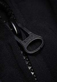 Černý jezdec zipu s vytištěným logem, připevněný k texturované tkanině s hladkým povrchem, s viditelným šitím podél okrajů.