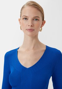 Niebieski prążkowany top z długim rękawem o dekolcie w kształcie litery V i teksturowanym wzorze. Modelka nosi złote kolczyki w formie kółek. Tło jest białe.