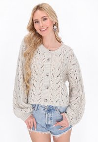 Šviesiai beige megzta kardiganas su nėriniuota raštu, pasižymintis didelėmis rankovėmis ir penkiomis didelėmis sagomis priekyje. Sukomplektuotas su džinsiniais šortais.