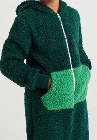 Pehmeä, hupullinen haalari tummanvihreänä, pehmeästä materiaalista; siinä on kirkkaanvihreä eri väriin kuuluva tasku ja valkoinen vetoketju. Hihansuut ja helma ovat ribbikangasta.