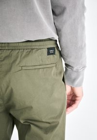 Pantalones verde oliva con una cintura elástica, bolsillo trasero y una etiqueta de logotipo negra. Acompañados de una camisa de manga larga gris.