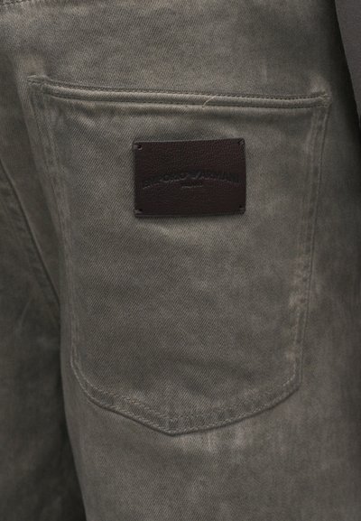 Tumed hallid püksid, millel on kitsas lõige, tekstuuriga kangas ja nahklogoga märgistus tagakoti peal, millel on reljeefne tekst.