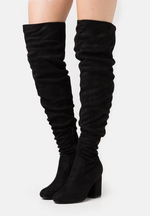Botas de ante negras por encima de la rodilla con tacones cuadrados y cremalleras laterales, llevadas sobre piernas al descubierto contra un fondo blanco.