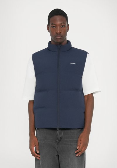 Marineblauwe puffervest met een hoge kraag en een ritssluiting aan de voorkant. Heeft een logo op de linkerborst en is gecombineerd met een wit shirt en grijze spijkerbroek.