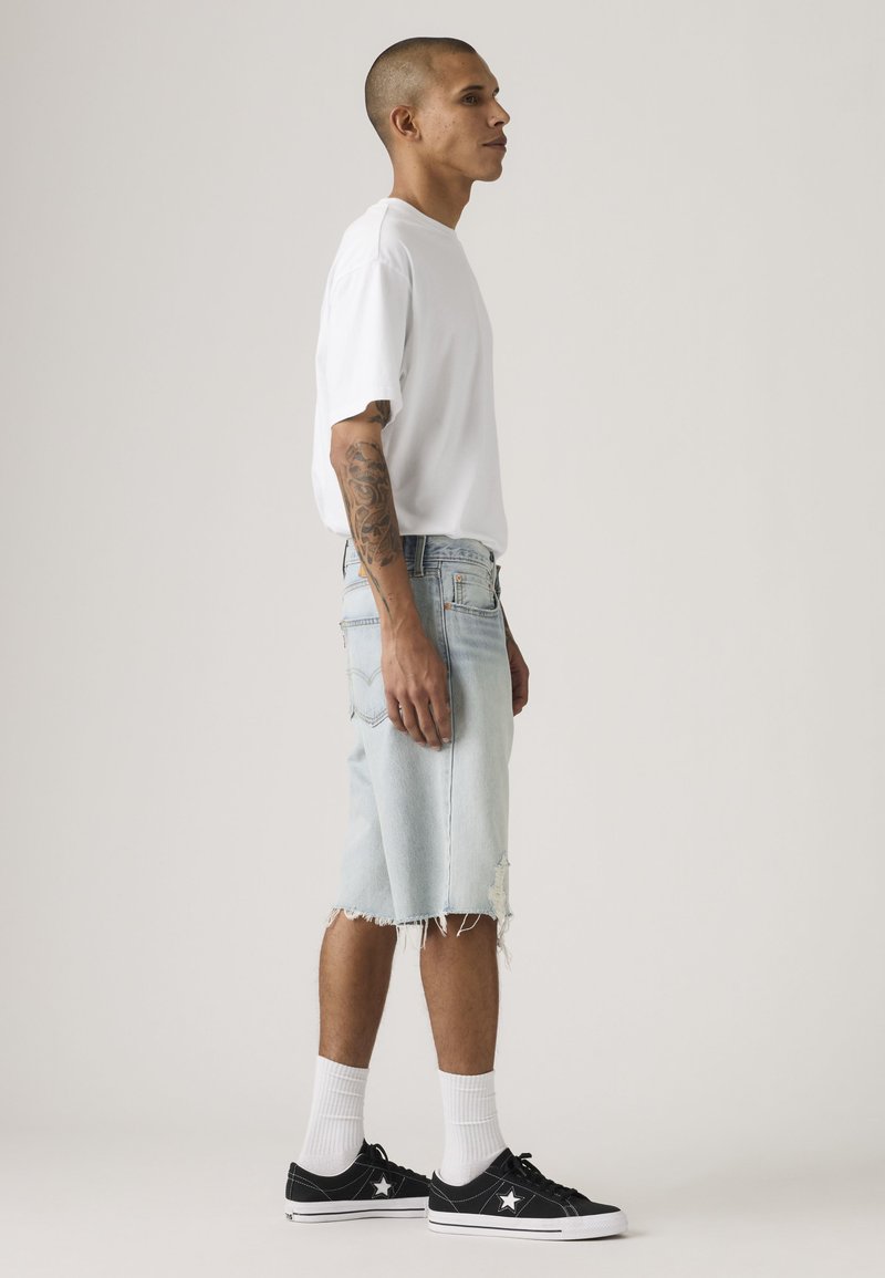 Jonge man met getatoeëerde arm draagt een wit T-shirt, licht gescheurde denim shorts, witte sokken en zwarte sneakers, staand in profiel.