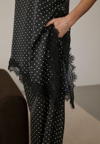 Main portant un bracelet en argent dans la poche d'un vêtement noir à pois avec bordure en dentelle noire, sur fond neutre près d'un meuble marron.