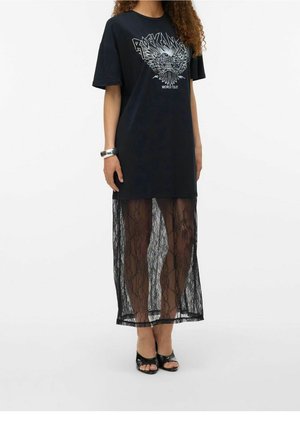 Kvinna iklädd svart grafisk t-shirtklänning med genomskinlig svart spetskjol ovanpå och svarta sandaler med öppen tå, stående mot vit bakgrund.