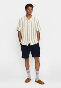 Camisa de manga corta, con collar, en un amarillo claro con rayas verticales azules, combinada con pantalones cortos azul marino y calcetines grises con rayas rojas y azules.
