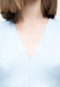 Γυναίκα με καστανά μαλλιά μέχρι τους ώμους που φοράει ανοιχτό μπλε μπλουζάκι με V λαιμόκοψη, εμφανιζόμενη από τους ώμους έως το μέσο του στήθους σε απλό φόντο.