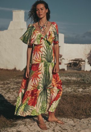 Woman with curly hair wears a colorful tropical-print dress and shell necklace, standing barefoot on grass near whitewashed buildings.