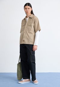 Beige kortärmad skjorta med två bröstfickor, svarta cargo-byxor med sidoficka, ljusrosa sneakers och olivgrön tote-väska.