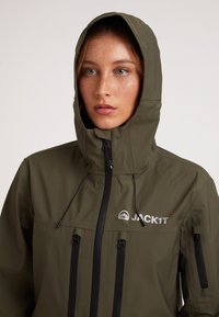 Giacca da esterno verde oliva con cappuccio, dotata di cerniere nere, logo sul petto e numerose tasche con zip per lo stoccaggio.
