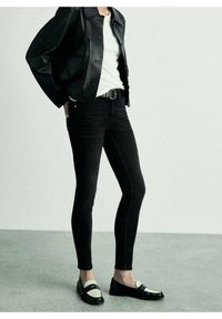 Vaqueros negros de diseño ajustado, combinados con una camisa blanca y una chaqueta de cuero negra. El conjunto incluye mocasines negros brillantes con detalles en blanco.