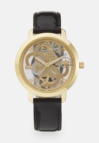 Orologio d'oro con quadrante trasparente che mostra ingranaggi intricati; cinturino in pelle nera lucida; grande design del logo luccicante.