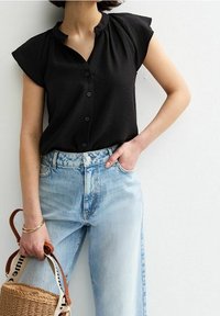 Blouse noire à manches courtes avec un devant boutonné et un col froncé, associée à un jean taille haute bleu clair et un sac en osier.