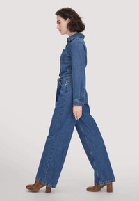 Blauer Denim-Jumpsuit mit langen Ärmeln, Kragen und weitem Bein. Strukturiertes Material mit Vordertaschen und Knopfdetails.