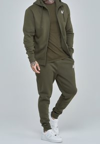 Conjunto de sudadera con capucha y pantalones de chándal en color verde olivo, hecho de tejido suave, con cremallera frontal, bolsillos y acentos de logo en blanco. Combinado con zapatillas blancas.