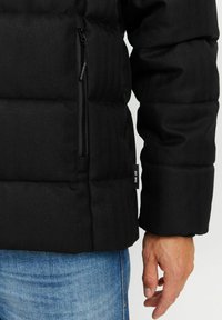 Schwarze Pufferjacke mit steppartigen Abschnitten, einer Reißverschlusstasche und einem kleinen Markenschild. Der Stoff verfügt über eine subtile Fischgrätenstruktur.