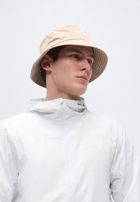 Νεαρός άντρας με μπεζ καπέλο τύπου bucket και ανοιχτό γκρι ζακέτα με φερμουάρ, στραμμένος προς τα δεξιά σε απλό λευκό φόντο.