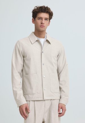 Junger Mann mit lockigem Haar trägt eine beige Jacke mit Knöpfen und zwei Vordertaschen über einer passenden beige Hose vor einem schlichten hellen Hintergrund.