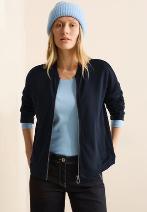 Giacca zip nera con polsini a coste, indossata sopra una camicia azzurra chiaro. Il modello indossa un berretto di maglia azzurra chiaro. Jeans scuri con cuciture visibili.