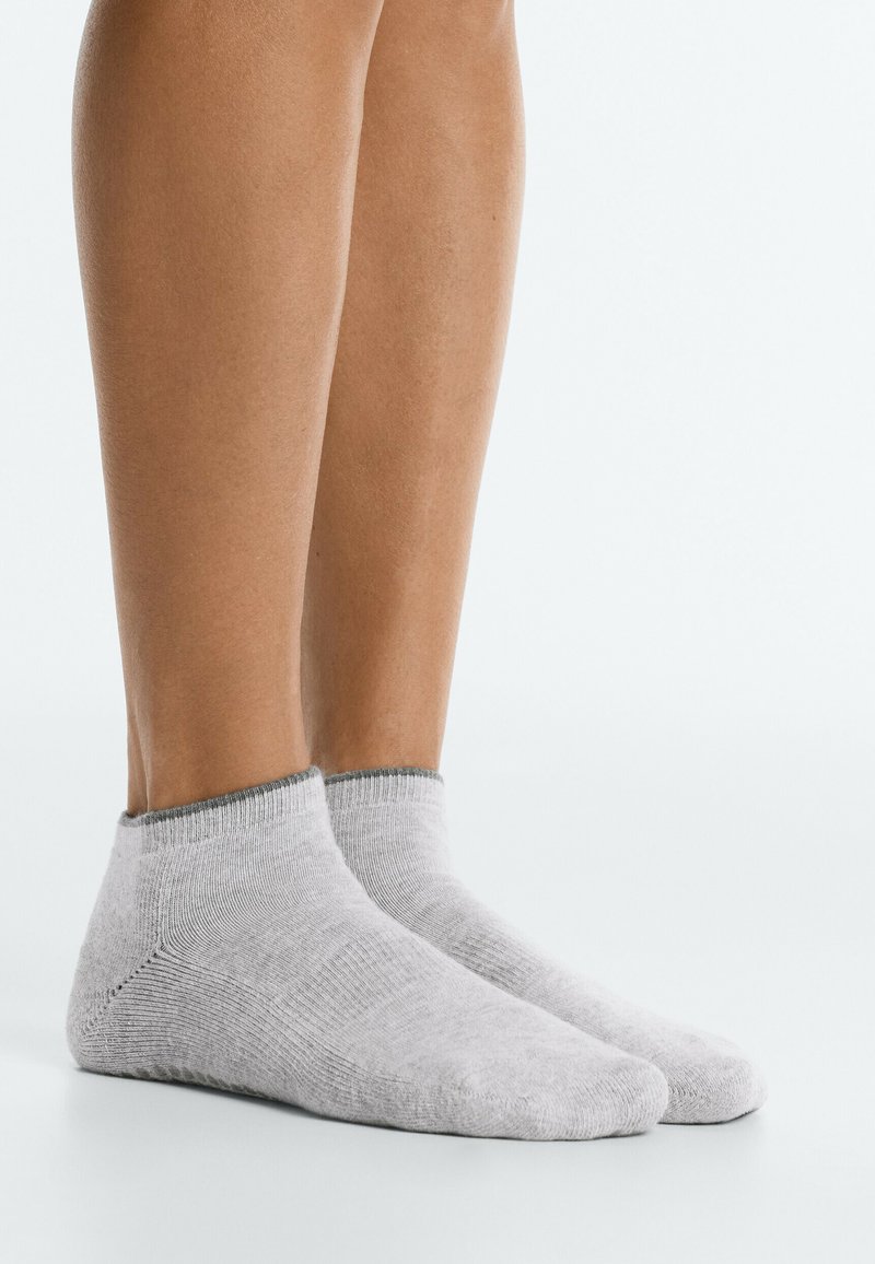 Paire de jambes portant des chaussettes courtes gris clair, debout sur une surface blanche avec un arrière-plan clair uni.