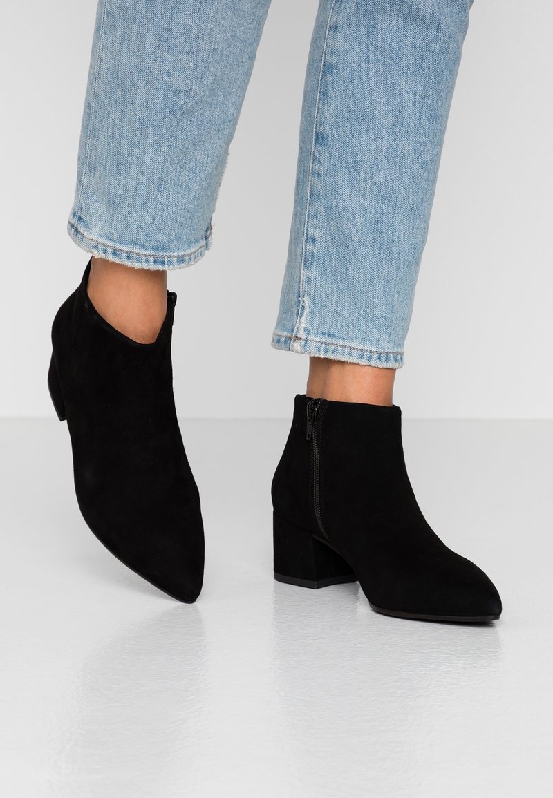 Bottines en daim noir à talons épais portées avec un jean court bleu clair sur fond blanc.