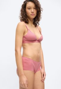 Νεαρή γυναίκα με κατσαρά μαλλιά που φοράει ροζ σουτιέν και αντίστοιχο σορτς, στέκεται μπροστά σε λευκό φόντο.