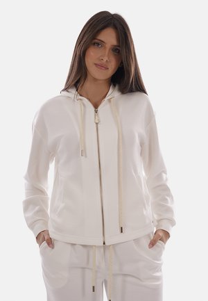 Donna che indossa una felpa bianca con cerniera e coulisse, abbinata a pantaloni bianchi, in piedi con le mani nelle tasche su uno sfondo semplice.