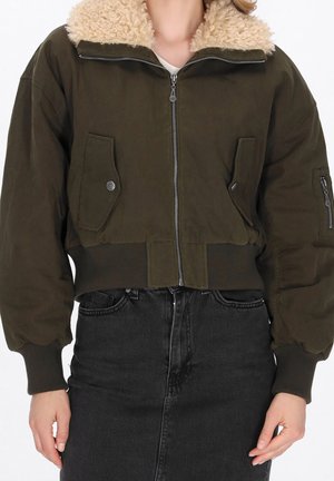 Vrouw draagt een donkergroene cropped bomberjas met schapenvachtkraag en zwarte high-waisted jeans, handen ontspannen langs het lichaam.