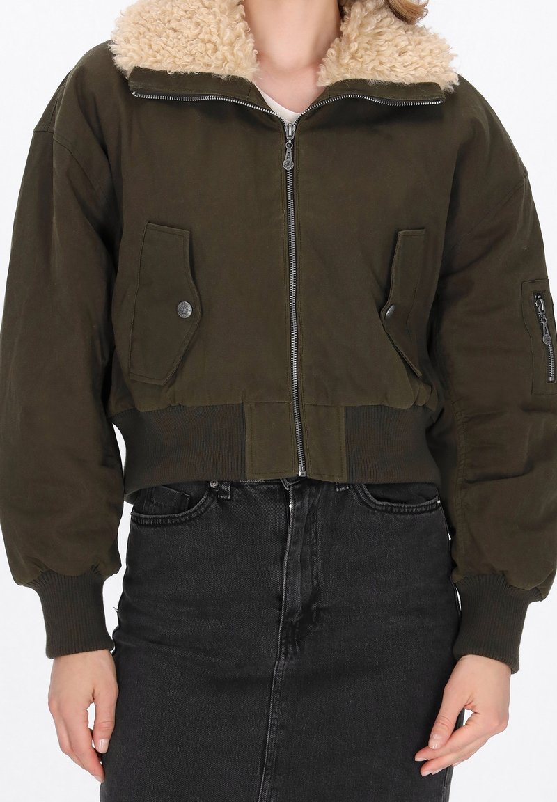 Femme portant une veste bomber courte vert foncé avec col en peau retournée et jean noir taille haute, mains détendues le long du corps.