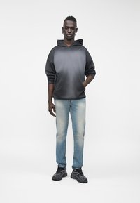 Hoodie com gradiente do preto ao cinza e bolso frontal, combinado com jeans de corte slim azul claro e ténis pretos. Fundo liso, sujeito centrado.