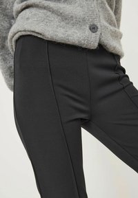 Pantalons noirs ajustés à la texture lisse, présentant une coupe slim et des coutures latérales. Accompagnés d'un cardigan en tricot doux de couleur gris clair.