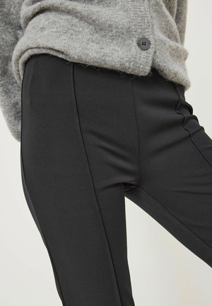 Schwarze, maßgeschneiderte Hose mit glatter Textur, im Slim-Fit-Stil und mit Seitennähten. Dazu wird eine hellgraue, weiche Strickjacke getragen.