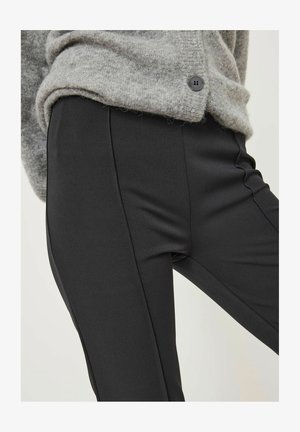 Pantalons noirs ajustés à la texture lisse, présentant une coupe slim et des coutures latérales. Accompagnés d'un cardigan en tricot doux de couleur gris clair.