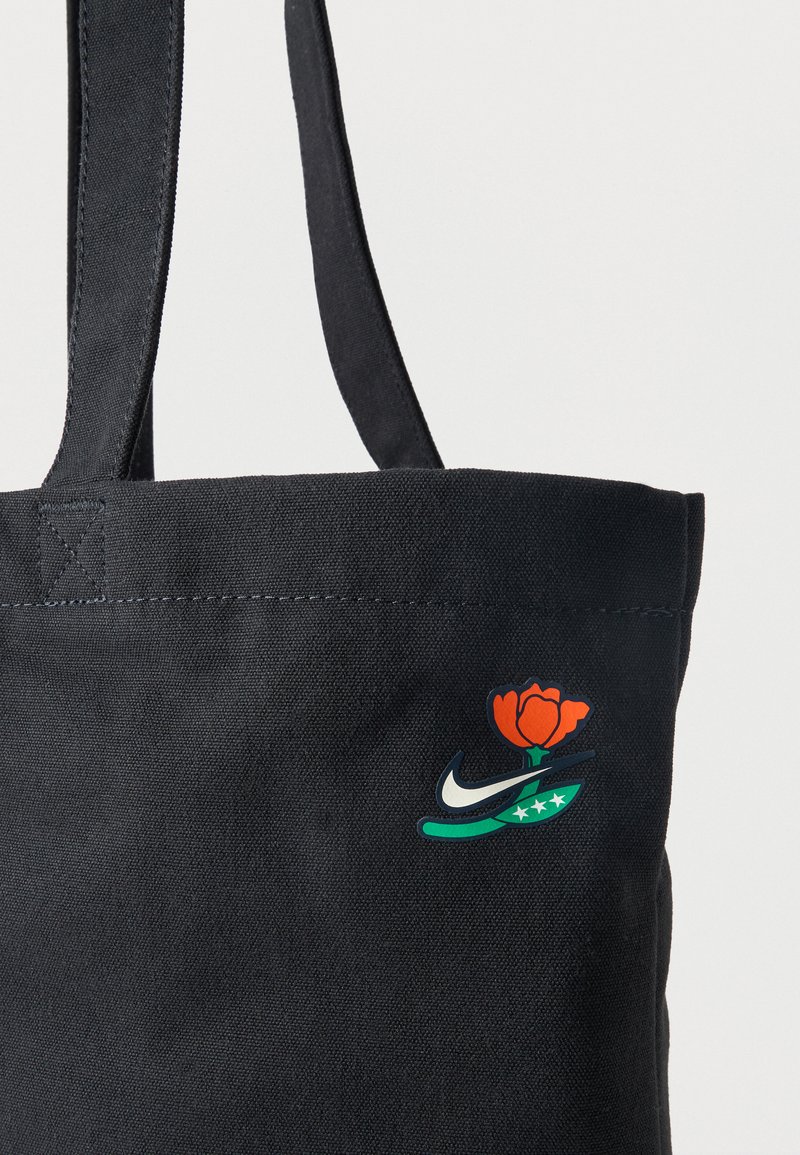 Bolsa tote de tela negra con dos asas y un pequeño logo de una rosa naranja, tallo verde con tres estrellas blancas y un swoosh blanco.