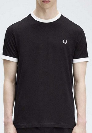 Hombre con camiseta negra de manga corta con cuello y bordes de las mangas blancos, pequeño logo de corona de laurel blanca en el pecho izquierdo.