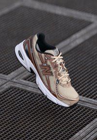 Baskets en mesh beige et marron avec une semelle rembourrée, comportant un logo "N" bien en vue, un col matelassé et un design à bout arrondi.