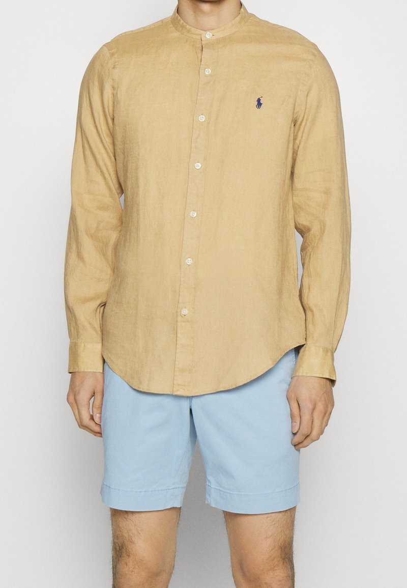 Světle hnědá košile s dlouhým rukávem a límcem, s knoflíkovým zapínáním vpředu a malým modrým logem na levé hrudi, spárovaná se světle modrými šortkami.