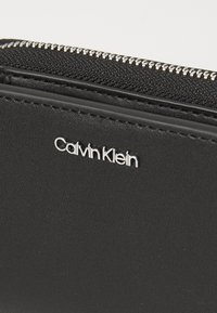 Svart läderplånbok med texturerad yta, utrustad med en silverfärgad dragkedja och präglad "Calvin Klein"-logotyp på framsidan.