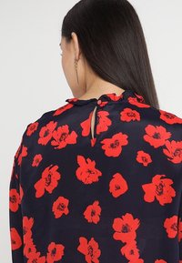 Marinblus med blommönster i röd färg, med en volangkrage och en nyckelhålsknäppning bak. Slarvigt, l ätt tyg.