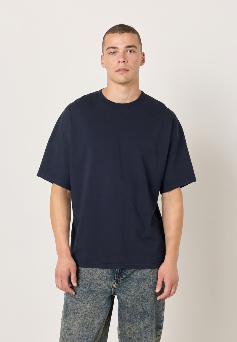 Temno modra bombažna majica s klasičnim okroglim izrezom in kratkimi rokavi, z rahlo ohlapnim krojem in gladko teksturo. Oblečena s svetlo pranim denimom.